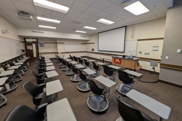 Front of room view with lectern on right in front of markerboard, projection screen down
