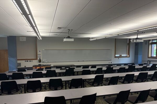 Back of room view of student tiered fixed-table and chair seating and exit doors at left and right rear of room
