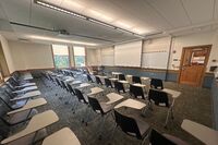 Front of room view with lectern on left in front of markerboard and projection screen lowered, exit door at front right of room