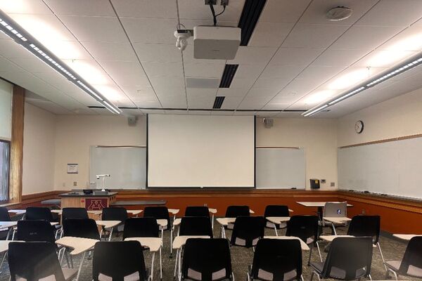 Front of room view with lectern on left in front of markerboard and projection screen lowered