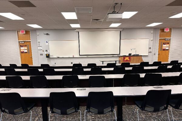 Front of room view with lectern on right in front of markerboard and projection screen lowered, exit doors to left and right