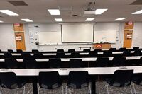 Front of room view with lectern on right in front of markerboard and projection screen lowered, exit doors to left and right