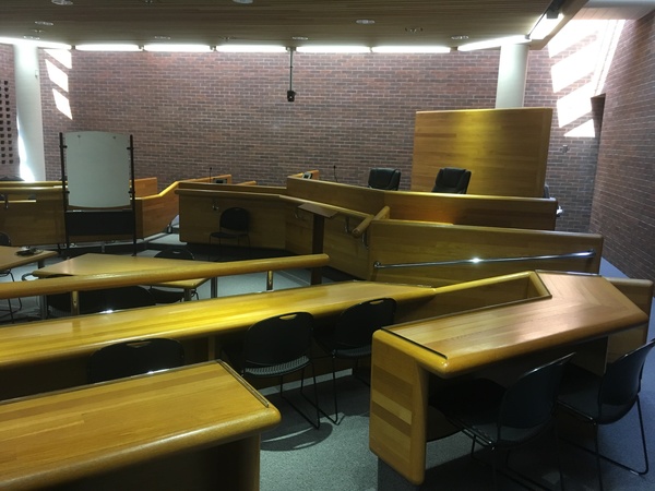 Photo of front of room from back of room showing a courtroom setup with door on the right, seating in half hexagon, and judges seating area.
