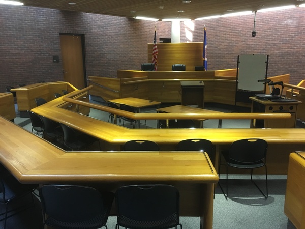 Photo of front of room from back of room showing a courtroom setup with door on the left, seating in half hexagon, and judges seating area.