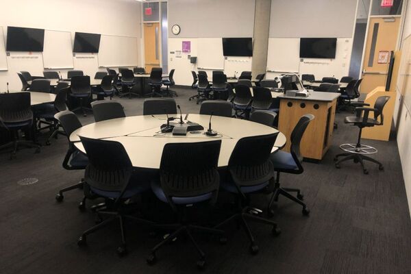 Room view of student active learning round table and chair seating, multiple markerboards and student display monitors on all walls, exit doors on left and right side of rear wall