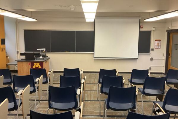 Front of room view with lectern on left in front of chalkboard, projection screen lowered