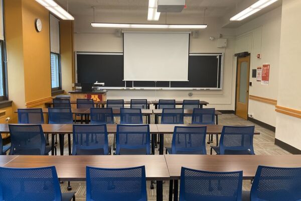 Front of room view with lectern on left in front of chalkboard, projection screen partially lowered 