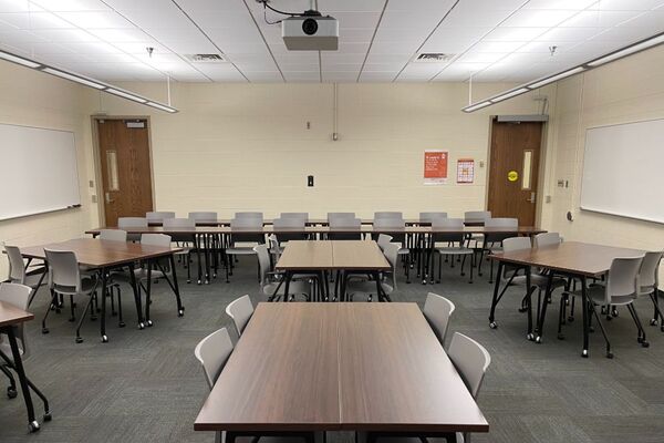 Back of room view of student table and chair seating, markerboards on right and left side walls, and exit doors at left and right rear of room
