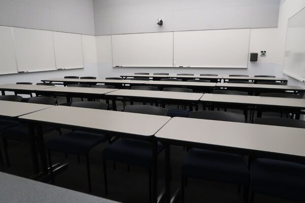 Back of room view of student table and chair seating, camera on rear wall, and markerboards on back and side walls of room