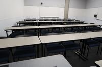 Back of room view of student table and chair seating, camera on rear wall, and markerboards on rear and right sidewall