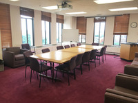 Photo of back of room from front of room with windows on front and left walls, conference room table in the middle, and couches along the perimeter.