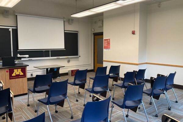 Front of room view with lectern on left in front of chalkboard and projection screen partially lowered