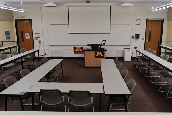 Front of room view with lectern center in front of markerboard and projection screen lowered, ext doors on left and right of markerboard