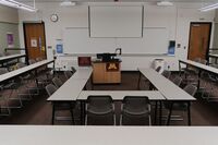 Front of room view with lectern center in front of markerboard and projection screen partially raised, exit doors on left and right of markerboard