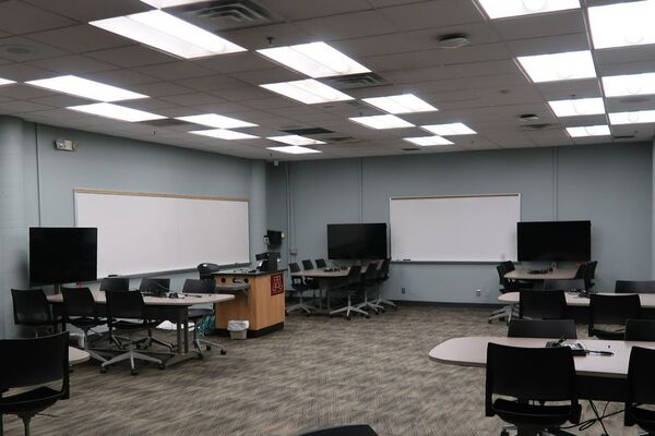 View of room view with lectern on left in front of markerboard, student group tables, and display monitors 