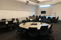 View with lectern on left in front of markerboard, markerboard and monitor on front wall, collaborative round tables and chair setting