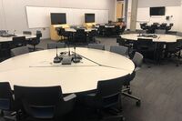 View of student active learning round table and chair seating, multiple markerboards and student display monitors on all walls, exit and lectern on middle right