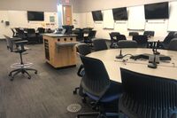 View of podium, student collaborative table and chair seating, display screens on side walls and exit door at rear of room