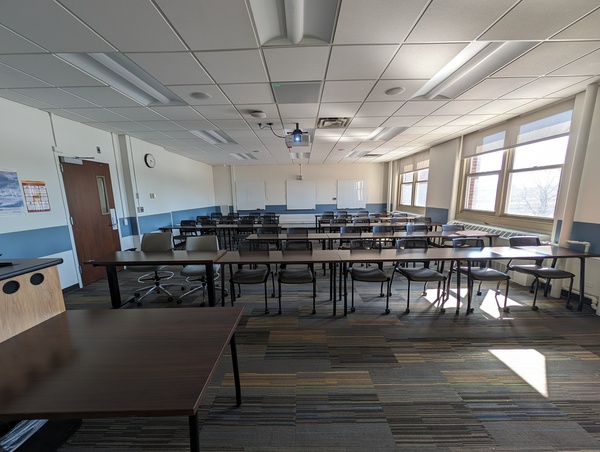 Back of room view of student table and chair seating and, camera and three markerboards on back wall of room