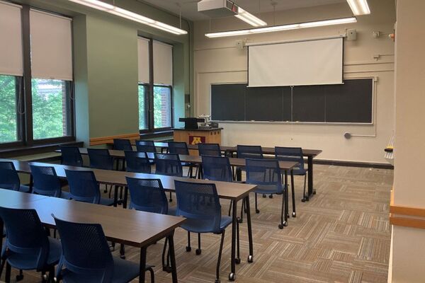 Front of room view with lectern on left in front of chalkboard and projection screen partially lowered