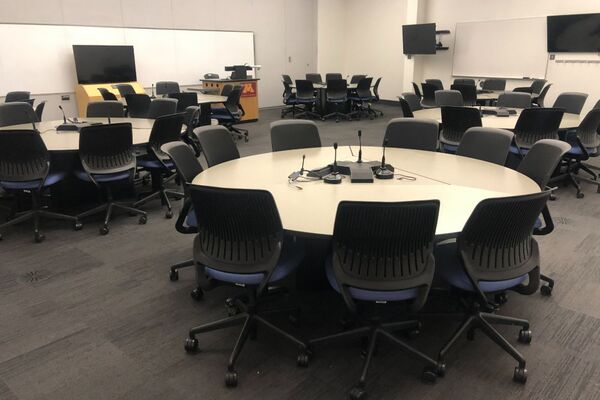 Room view of student active learning round table and chair seating, multiple markerboards and student display monitors on all walls, lectern on middle left