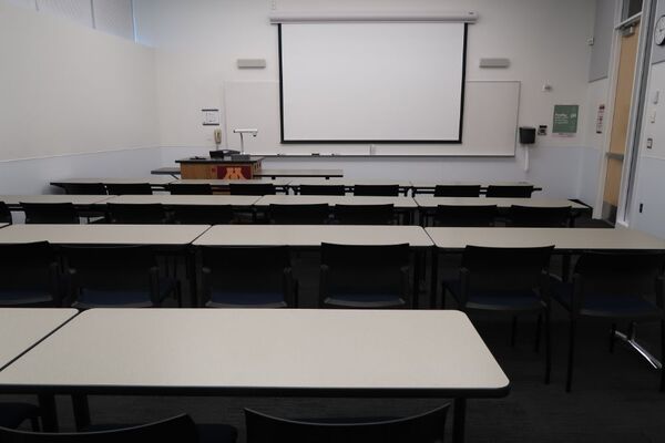Front of room view with lectern on right in front of markerboard and projection lowered, exit door on right side wall