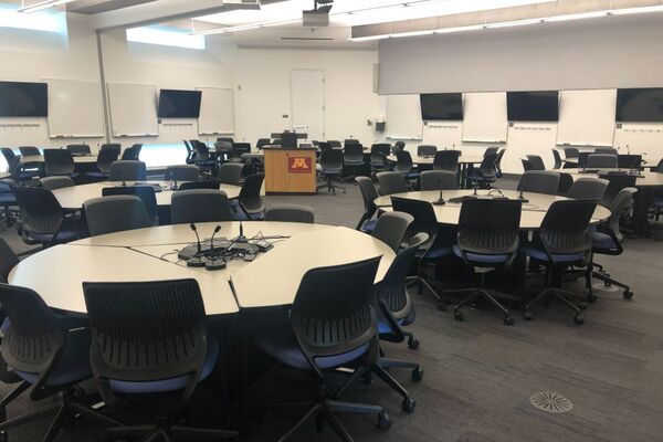 View of student collaborative table and chair seating. Lectern is in center-back of room in front of monitors and markerboards on both side walls.