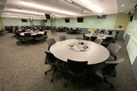 Room view of student active learning table seating, markerboards on all walls, and two exit doors at right side of room