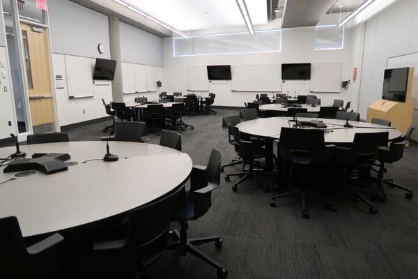 Room view of student active learning round table and chair seating, multiple markerboards and student display monitors on all walls, exit door on left wall