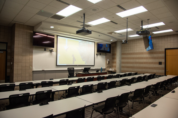 Audience view from rear left.  Two displays next to the front projector display behind the podium are visible.  Room camera hanging from the ceiling can be seen