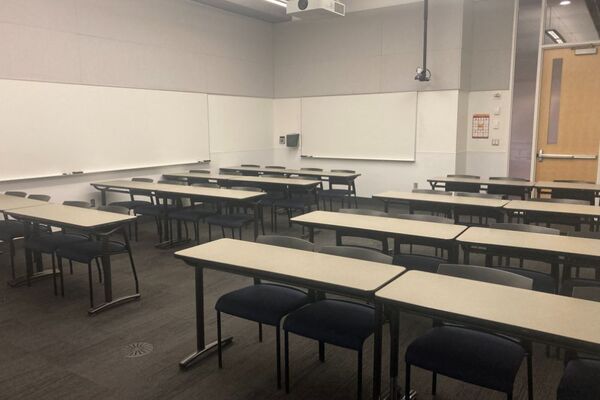 Back of room view of student table and chair seating, markerboards on left and rear wall, and exit door at right rear of room