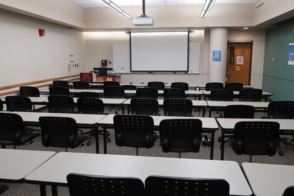 Front of room view with lectern on left in front of markerboard and projection screen lowered, exit door on front right