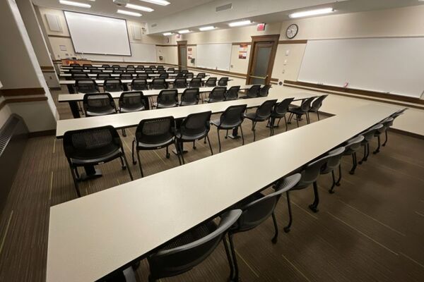 Front of room view with lectern on left in front of markerboard and projection screen lowered, exit door on front right wall