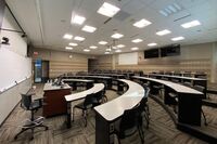 Back of room view of student tiered fixed-table and chair seating and projection screen center, confidence monitor on the right