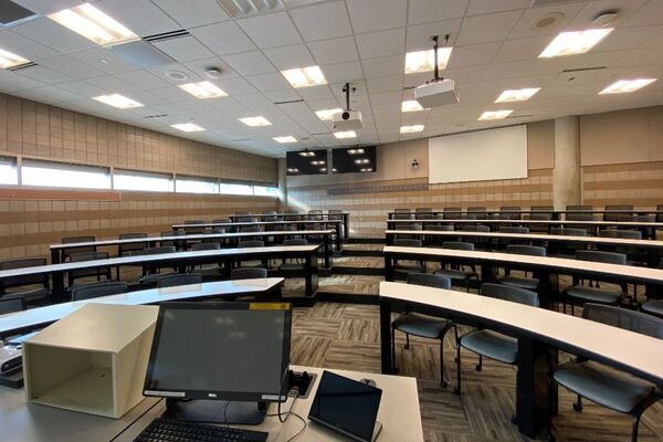 Back of room view of student tiered fixed-table and chair seating, project screen and confidence monitor on rear wall
