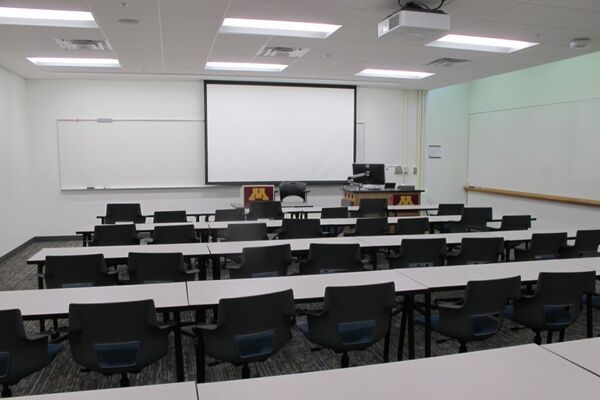 Front of room view with lectern on right in front of markerboard and projection screen lowered
