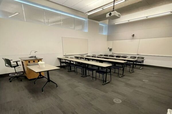 Back of room view of student table and chair seating, camera on rear wall, and markerboards on rear and right side wall