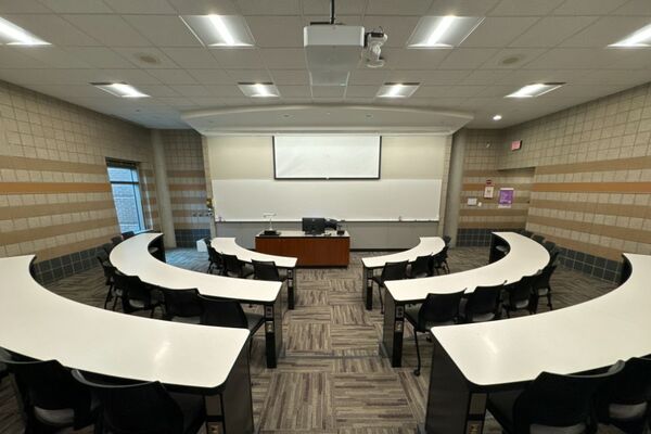 Front of room view with lectern center in front of markerboard, exit door on front right, screen partially lowered