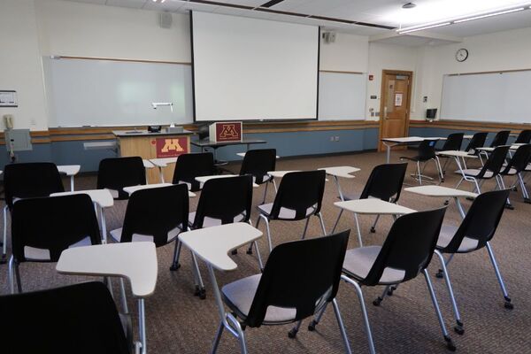 Front of room view with lectern on left in front of markerboard and projection screen lowered, exit door at front right of room