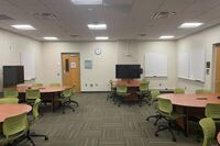View of student table and chair seating, student table displays, markerboards, and exit door on the left