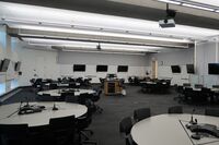 Room view with lectern in center of room, markerboards and student display monitors are on all walls, exit door on right wall 