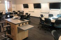 View of student collaborative table and chair seating, markerboards and display screens on wall, and exit door on left of room