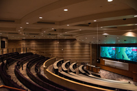 Audience view of entire room from rear right.  The majority of audience seating is visible, along with student mics hanging from ceiling, front podium, and projector displays/whiteboard.