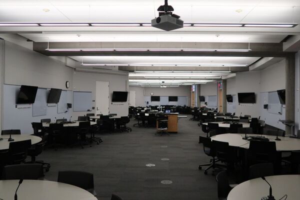Room view with lectern in center of room, walls of room have multiple markerboards and student display monitors, three exit doors on right wall of room