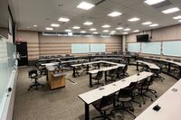 Back of room view of student tiered fixed-table and chair seating, markerboards on back and side walls, monitor on back wall