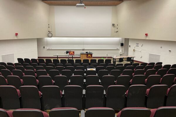 Front of room view with lectern on right in front of markerboard and projection area above, exit door center right