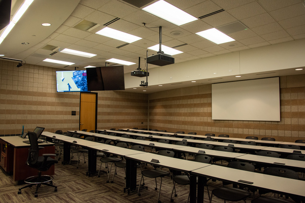 Instructor view from front right of classroom showing two confidence panels hanging from ceiling, rear projector screen at back of classroom, student seating in rows, and room camera.  Front podium is at the far left of the image.
