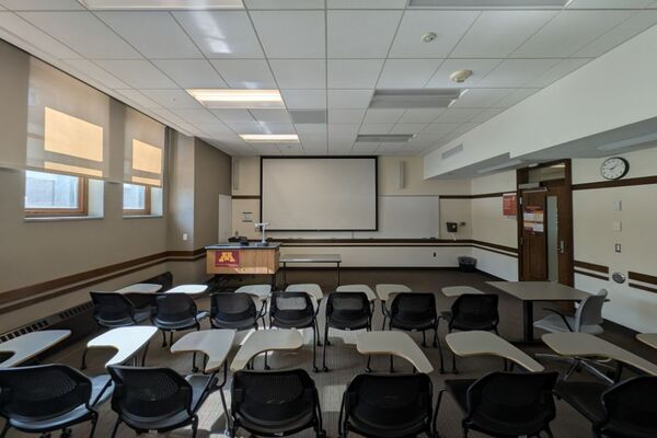 Front of room view with lectern on left in front of markerboard and projection screen lowered