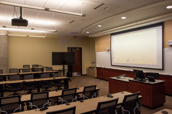 View from audience right side showing instructor confidence monitor, instructor podium, project and projector display, front board, student microphones and seating, and exit door
