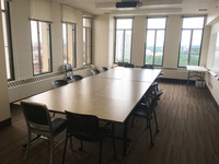 Photo of back of room from front of room, windows on the left and back walls, seating set like a conference room table.
